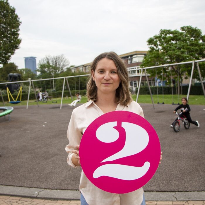 Michelle is procesmanager Wijkaanpak staat bij een kinderspeelplaats in de Schilderswijk in Den Haag waar zij zelf ook heeft gewoond. Ze heeft een groot cijfer 2 in haar handen, het logo van 2Samen.