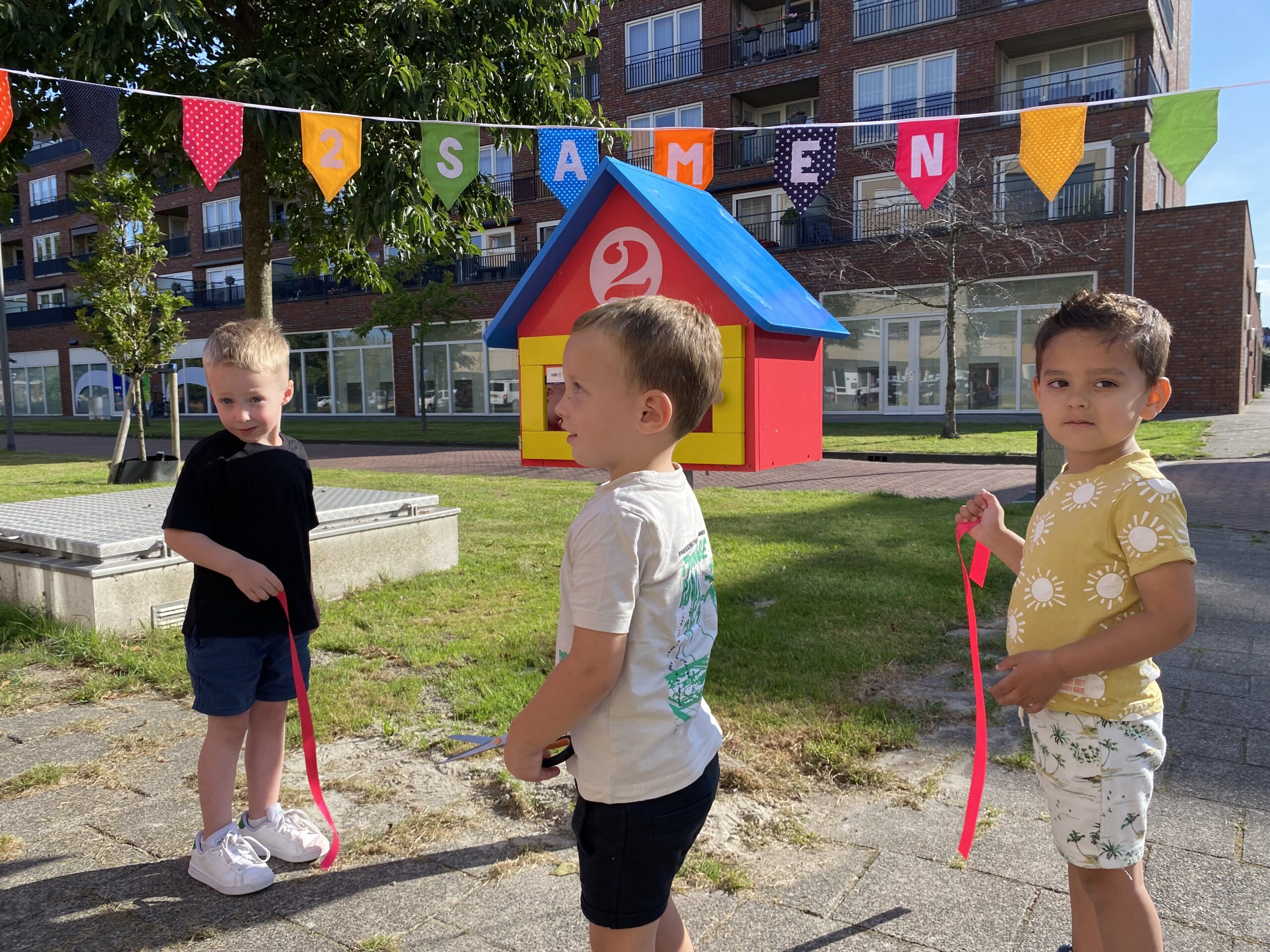 Het lint is doorgeknipt dus de mini-bieb is geopend bij kvd 2Eekhoorns in Hoogeland Naaldwijk. Drie peuters kijken blij en trots